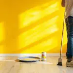 A homeowner standing in a cleared room with a paintbrush and paint can on the floor, preparing for an interior house painting project.